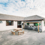 A welcoming, detached single-story bungalow under a bright sky, featuring a spacious paved courtyard with an outdoor picnic table. A sheltered storage area holds bicycles and a green kayak, signaling an active, youthful, and secure home environment managed by Green Light